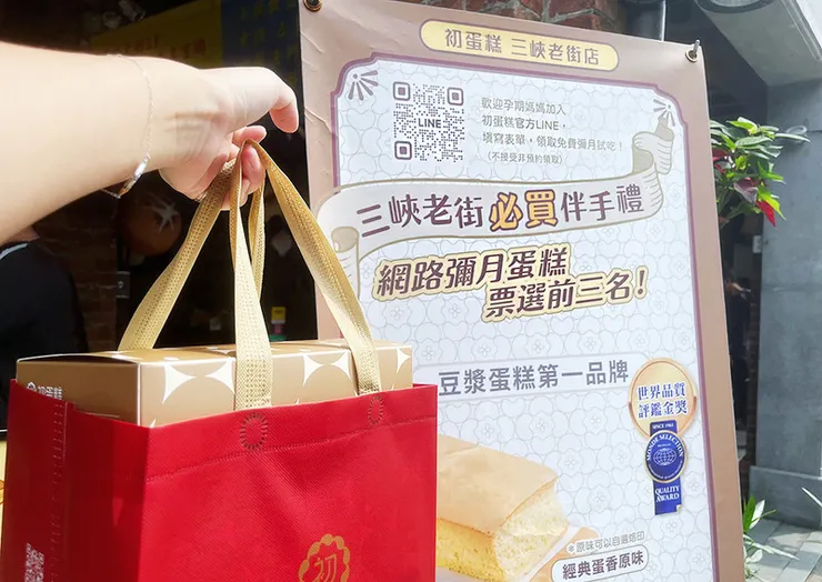 提紅色品牌購物袋，裝有金黃色系禮盒，背景為「初蛋糕三峽老街店」布條，標榜網路票選彌月蛋糕前三名與豆漿蛋糕第一品牌，適合送禮情境。