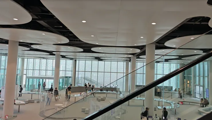 台中綠美圖淺色調地面與白色圓柱支撐起挑高層級，自動扶梯與開放式閱讀區並列，讀者穿梭其間，營造出舒適的公共閱讀氛圍。，構築自然共生的現代閱讀場域