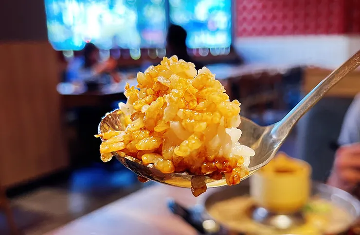 銀色湯匙上盛滿了拌好深色豬油醬汁的米飯，看起來晶瑩剔透，醬色濃郁，背景是模糊的餐廳場景。