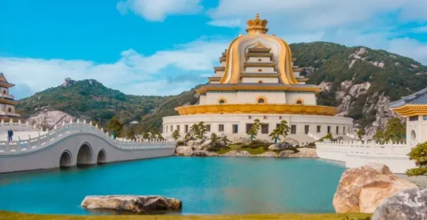 圖片顯示了一座宏偉的觀音寺廟,寺廟的金色屋頂與周圍的湖泊和綠意盎然的景色相互映襯。畫面中還有一座拱橋橫跨湖面,增添了景點的壯麗與寧靜感。