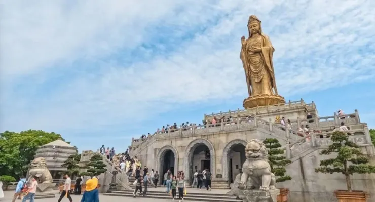 南海觀音立像,高聳於山頂,周圍人群攀登階梯前往敬拜,金色的觀音像在藍天的映襯下顯得格外莊嚴。