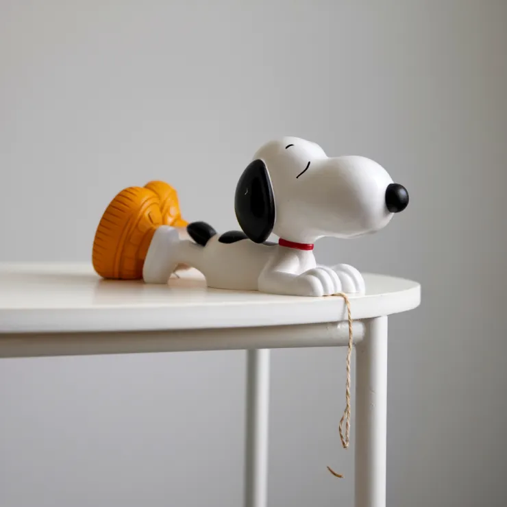 A photo of a plastic Snoopy figure sculpture placed on a white table. Snoopy is lying down with his back arched, his head peeks out from the edge of the table, and he holds a string that hangs over the edge of the table. The background is white.