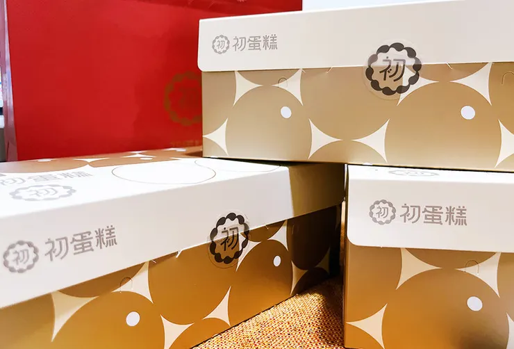 三款堆疊的初蛋糕品牌禮盒特寫，白底配上金黃色幾何圖案設計，印有「初蛋糕」Logo 與「初」字圓形貼紙，視覺簡約大方且具質感。