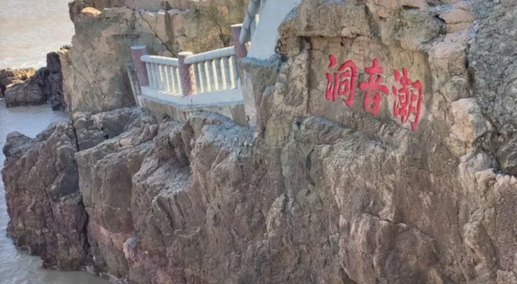 潮音洞入口處的岩石上刻著「潮音潮」的紅色字樣,旁邊有一條石階小徑通向洞口,背景是湛藍的海水和岩石。