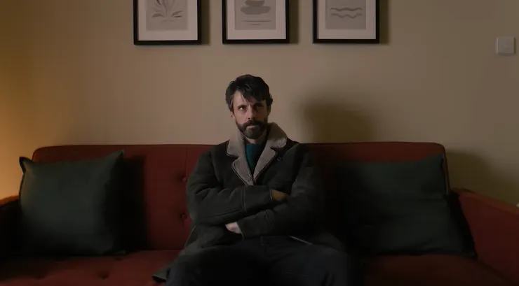 坐在沙發上，雙手交叉、神情警戒——那是他的防衛姿勢，也是一種內心的掙扎。