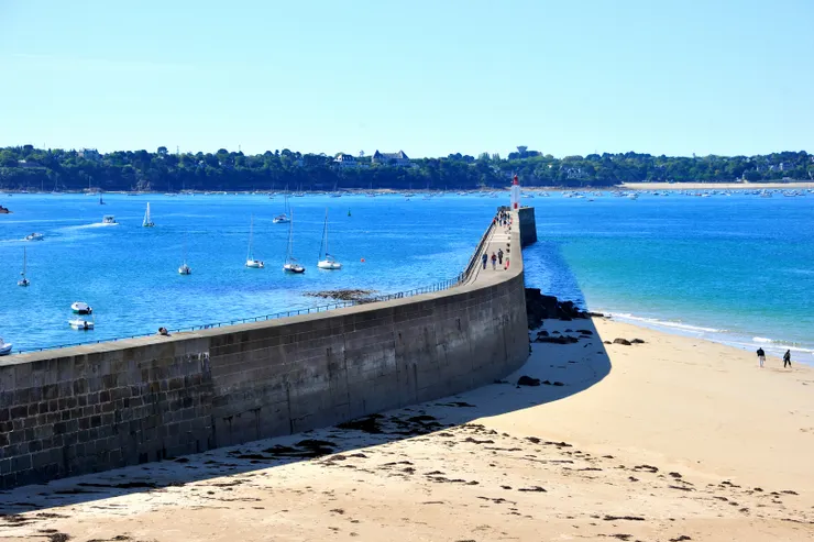 【城牆俯瞰，一條長堤延伸入海，盡頭有一座燈塔，堤防上有人們在散步，海灣中泊有許多帆船，顯示港口城市特色。】