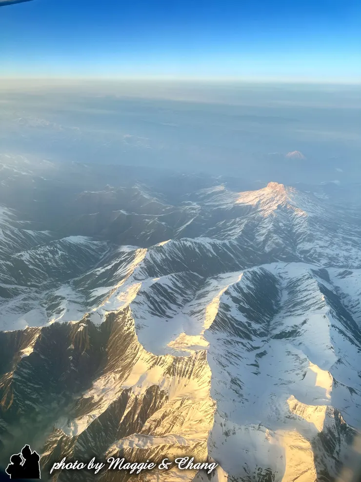 陽光輕灑，山峰間的陰影層次分明，展現出一種雄偉、壯麗又帶點神秘的氛圍。遠方的天際線由深藍漸變為淡藍，與薄霧交錯，宛如天地相接的夢幻邊界。