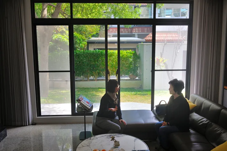 【上桌前兩位女客在客廳聊天，落地窗外，花木扶疏，樟樹提供大片綠意與蔽蔭。】