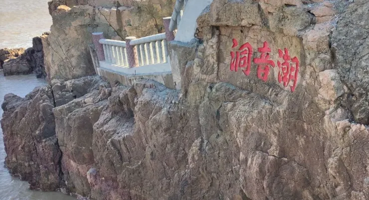 潮音洞入口處的岩石上刻著「潮音潮」的紅色字樣,旁邊有一條石階小徑通向洞口,背景是湛藍的海水和岩石。