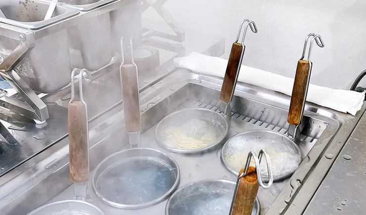 白拉麵廚房煮麵台特寫，煮麵網內正在沸水中烹煮細麵，熱氣騰騰展現日式拉麵精準火候與職人精神。