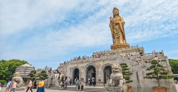 南海觀音立像,高聳於山頂,周圍人群攀登階梯前往敬拜,金色的觀音像在藍天的映襯下顯得格外莊嚴。