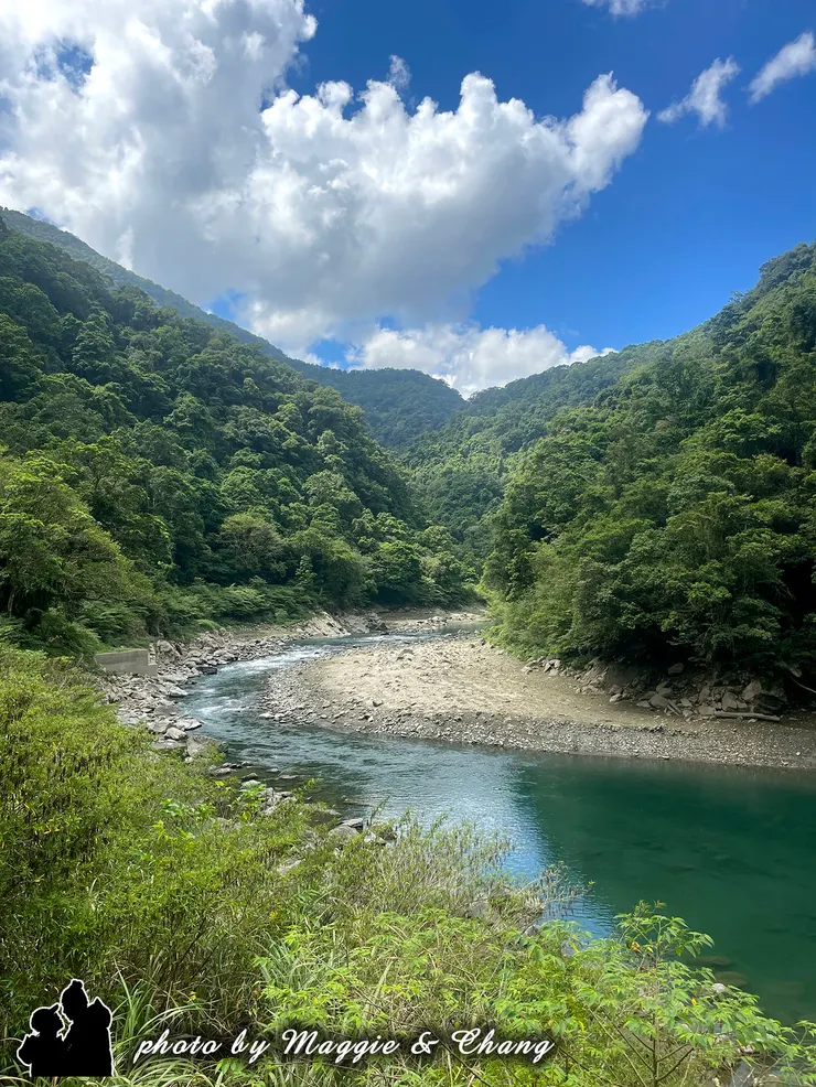 藍天白雲映照在翠綠山巒之上，蜿蜒的溪水靜靜流淌，水面呈現碧綠與深藍的漸層色澤，彷彿一幅大自然的油畫。這裡空氣清新，讓人感受到遠離塵囂的寧靜。
