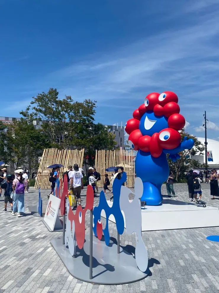 萬博吉祥物名字叫「ミャクミャク（脈脈）」，造型是紅白藍三色搭配，全身像是液態血管的集合體，頂著一顆充滿眼睛的頭。據官方解釋，他象徵生命的脈動與連結，體現萬博主題「設計未來社會，想像明日生活」。