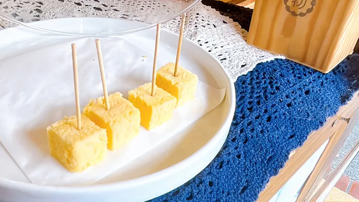 白色圓盤上整齊排列著四塊正方形豆漿蛋糕試吃品，插著牙籤供遊客品嚐，呈現出蛋糕天然的金黃色澤與孔洞均勻的鬆軟質地。