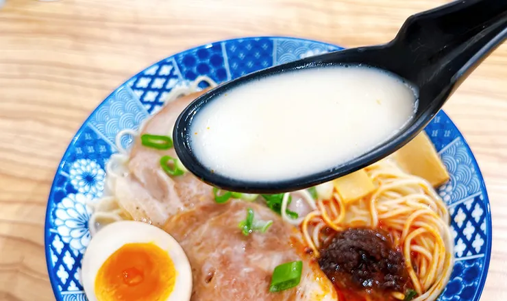 湯匙舀起小白拉麵的麻辣豚骨湯頭，展現未拌開前純白濃郁的豚骨原湯與特製麻辣紅油的強烈對比。