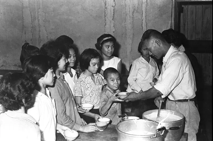 美援食物。小朋友們紛紛拿著碗排隊，準備領取大人用湯匙舀著鍋子內所煮好的食物。（圖片來源：國家文化記憶庫）