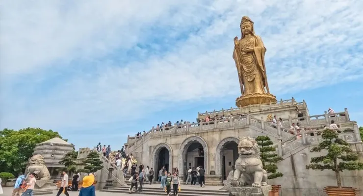 南海觀音立像,高聳於山頂,周圍人群攀登階梯前往敬拜,金色的觀音像在藍天的映襯下顯得格外莊嚴。