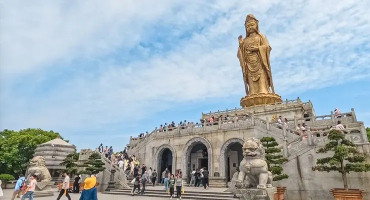 南海觀音立像，高聳於山頂，周圍人群攀登階梯前往敬拜，金色的觀音像在藍天的映襯下顯得格外莊嚴。