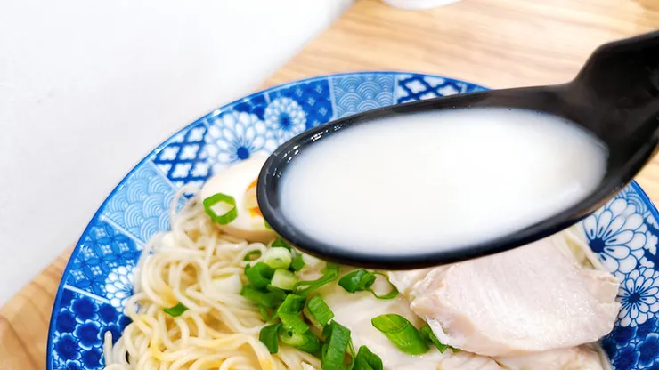 湯匙舀起小白拉麵的雞豚白湯，湯色呈現天然乳化後的純白色澤，富含膠質與純粹的熬煮精華。