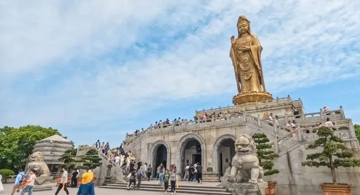 南海觀音立像，高聳於山頂，周圍人群攀登階梯前往敬拜，金色的觀音像在藍天的映襯下顯得格外莊嚴。