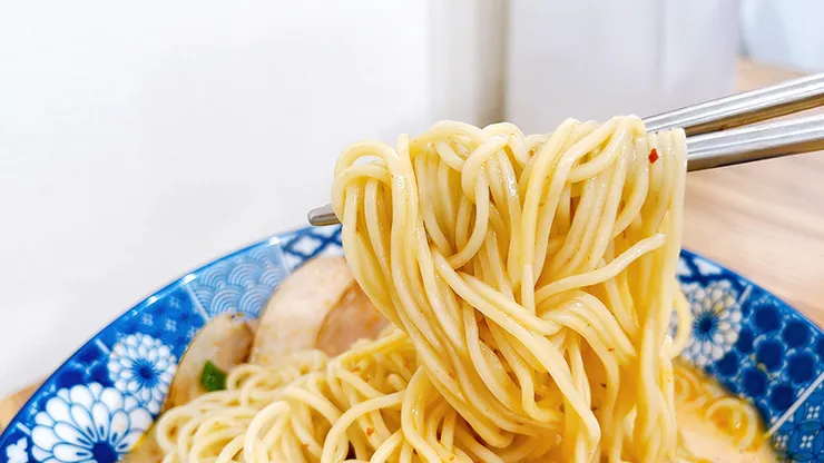 筷子夾起小白拉麵的日式細麵，麵條吸附滿滿的麻辣豚骨紅油湯汁，展現Q彈誘人的拉麵特寫。