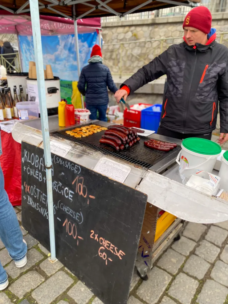 ▲捷克街頭小吃～烤香腸（Klobása）——那種煙燻香腸，色澤深而亮，味道馥郁而各家私房香料功夫皆不同。麵包夾烤香腸加上芥末醬，一份120克朗。特別的是，煎台上那黃色的「煎起司」，使用的起司是斯洛伐克生產的煙燻羊奶起司（Oštiepok），4片煎起司搭配蔓越莓醬，這樣一份100克朗。