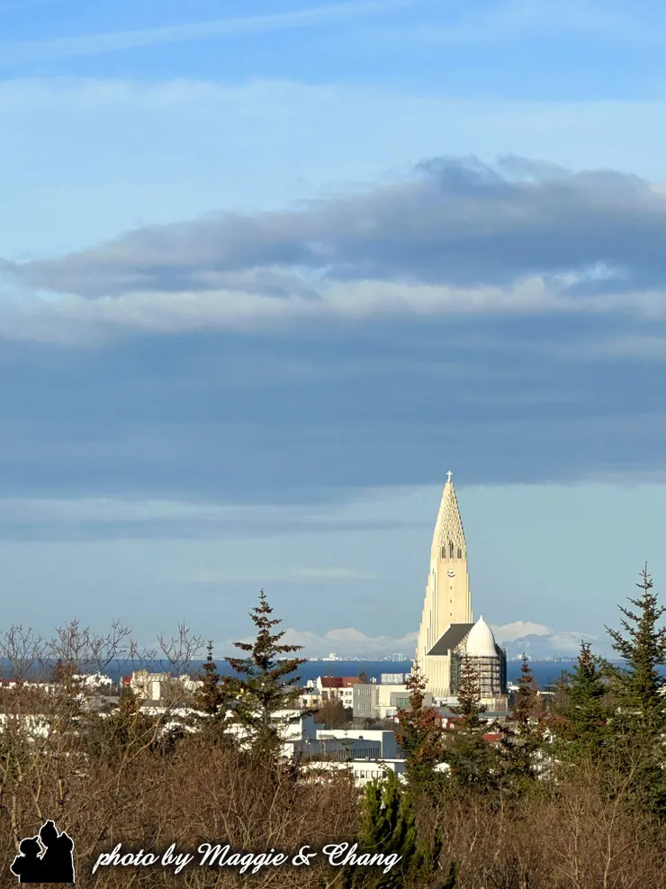 哈爾格林姆教堂塔樓筆直聳立於市區之中。前景是冬季枯木與松樹,遠方可見積雪覆蓋的山脈,藍天與層層雲層映襯出寧靜又壯闊的北國景致。