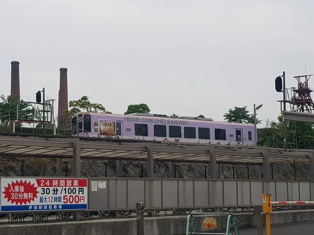 正從第二月台出站的平成筑豐鐵道列車，背景是田川市石炭公園內的兩根大煙囪