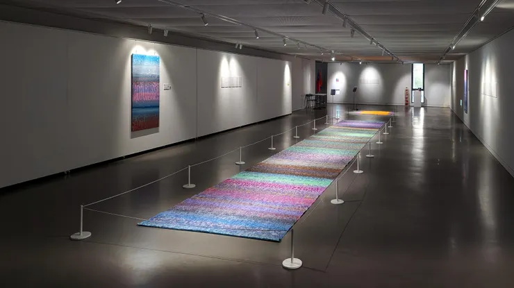 積吶虹光〈青天白日滿地虹：潮間帶的對話〉2018年，壓克力顏料、宣紙，1255×188 cm。色彩就像一條無止盡的路，在每一條垂直線交錯的瞬間，從寧靜到喧囂，從夢幻到現實。畫也不只是畫，而是心靈的回聲，在無邊的天地間，無聲歌頌著那跨越種族與地域的《普世契約，青天白日滿地虹》。這件鋪設於地面的作品透過觀眾的『行走』實踐『身體化』倫理，強調這份契約必須紮根於『此時此地』的共 同行動，而非寄託於虛無的超驗承諾。