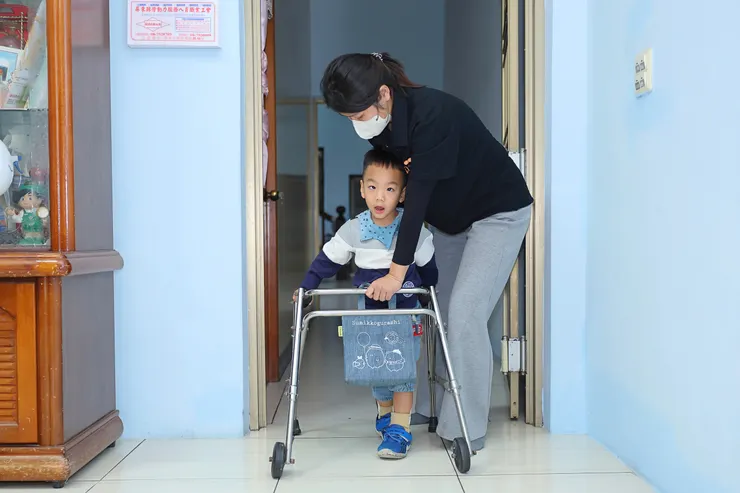 （圖說：經過半年的練習，睿睿在教保員徐鈺喬協助下，練習用助行器向前移動。）